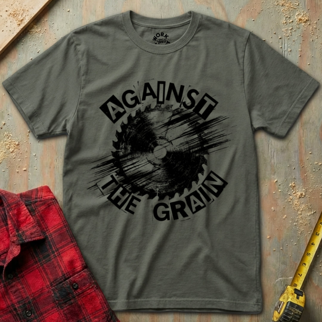 Gray t-shirt with 'Against the Grain' graphic design on a textured surface with a red plaid shirt and yellow tool in the background.