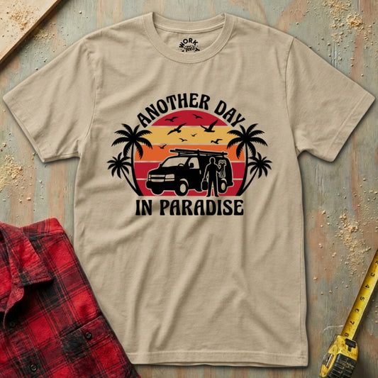 Beige t-shirt with 'Another Day in Paradise' graphic design on a wooden surface with a red plaid shirt and yellow tool in the background.
