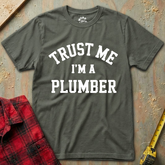 Gray t-shirt with 'Trust Me I'm a Plumber' text on a textured surface with a red plaid shirt and yellow tool in the background.