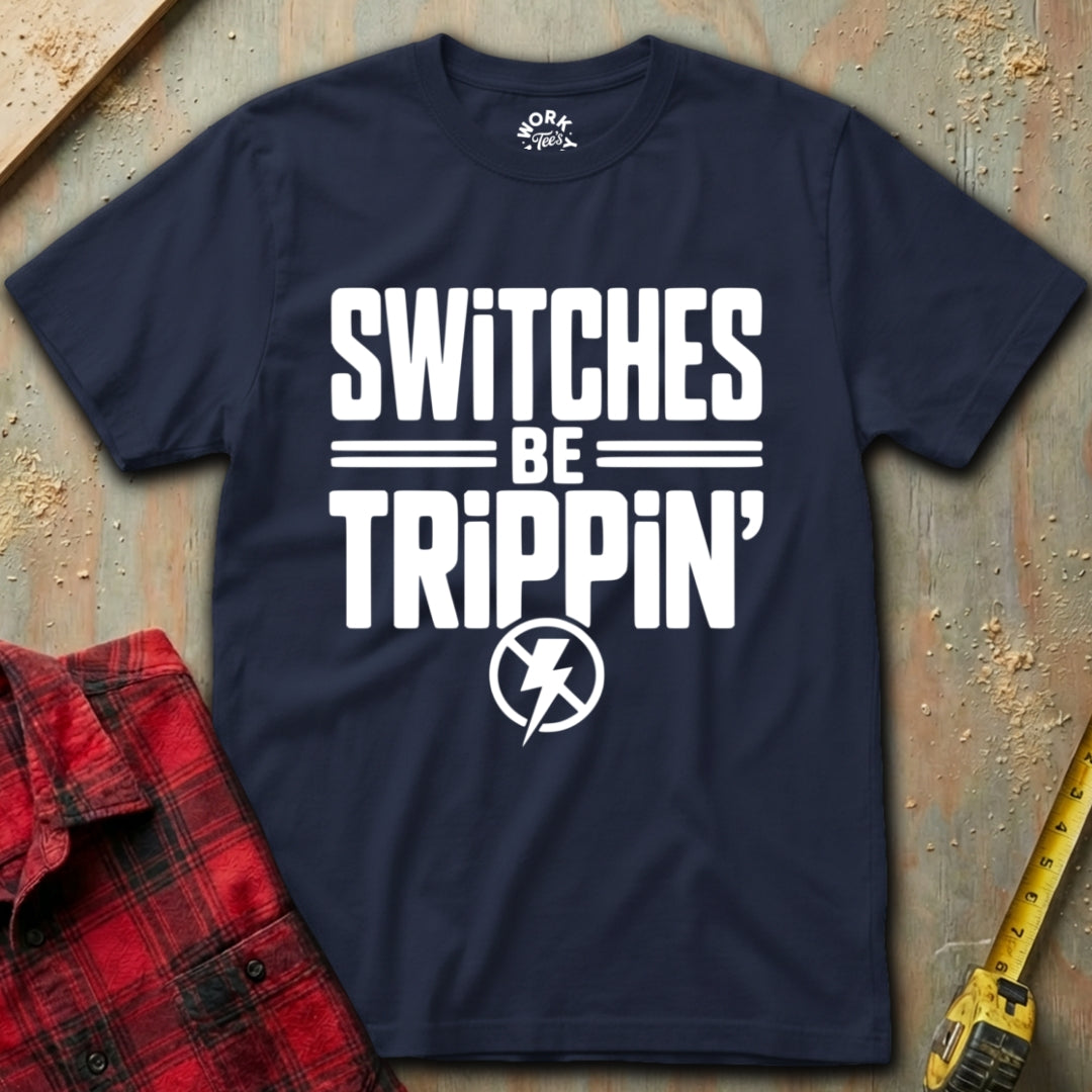 Navy blue t-shirt with white text 'SWITCHES BE TRIPPIN'' on a textured surface with a red plaid shirt and yellow tool in the background.