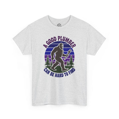 White t-shirt with a graphic of a bigfoot-like creature holding a wrench, surrounded by trees, with text 'A good plumber can be hard to find' on a white background.