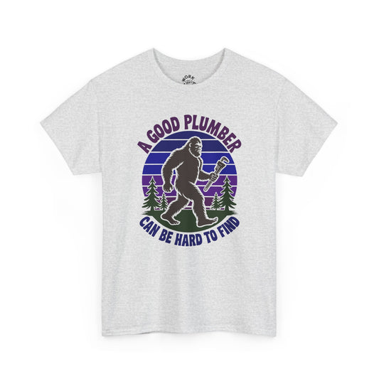 White t-shirt with a graphic of a bigfoot-like creature holding a wrench, surrounded by trees, with text 'A good plumber can be hard to find' on a white background.