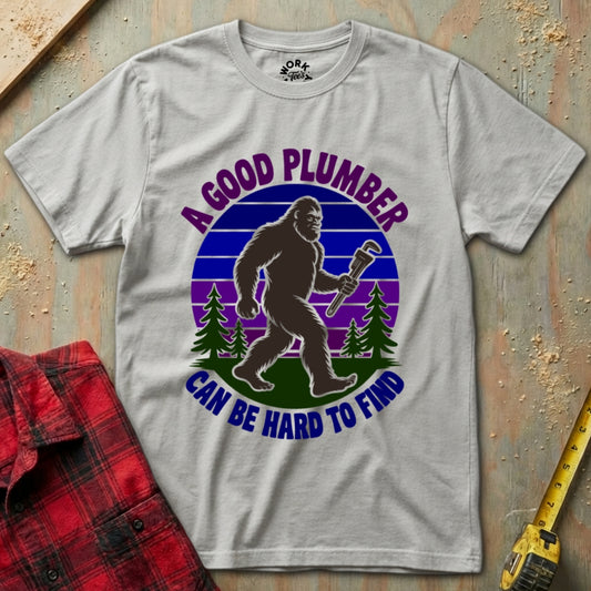 Gray t-shirt with a graphic of a bigfoot holding a wrench and text 'A good plumber can be hard to find' on a textured surface.