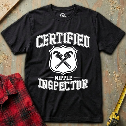 Black t-shirt with 'Certified Nipple Inspector' graphic on a textured surface with a red plaid shirt and yellow tool in the background.