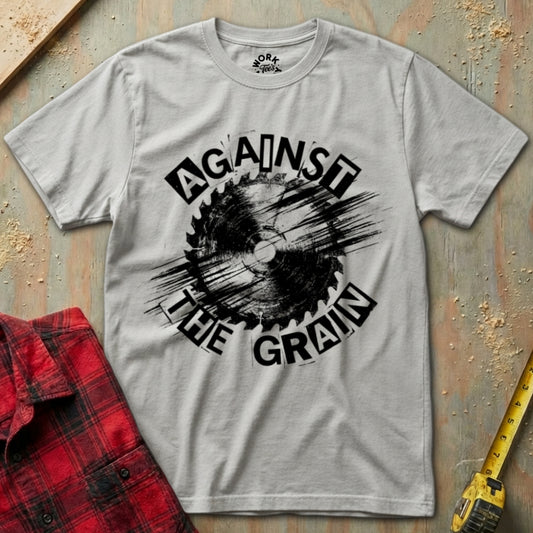 Gray t-shirt with 'Against the Grain' graphic design on a textured surface with a red plaid shirt and yellow tool in the background.