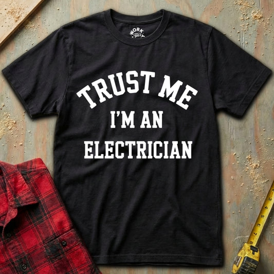 Black t-shirt with 'Trust Me I'm an Electrician' text on a textured surface with a red plaid shirt and yellow tool in the background.