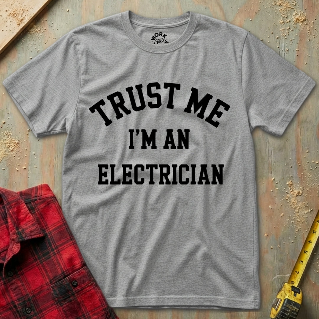 Gray t-shirt with 'Trust Me I'm an Electrician' text on a textured surface with a red plaid shirt and yellow tool in the background.