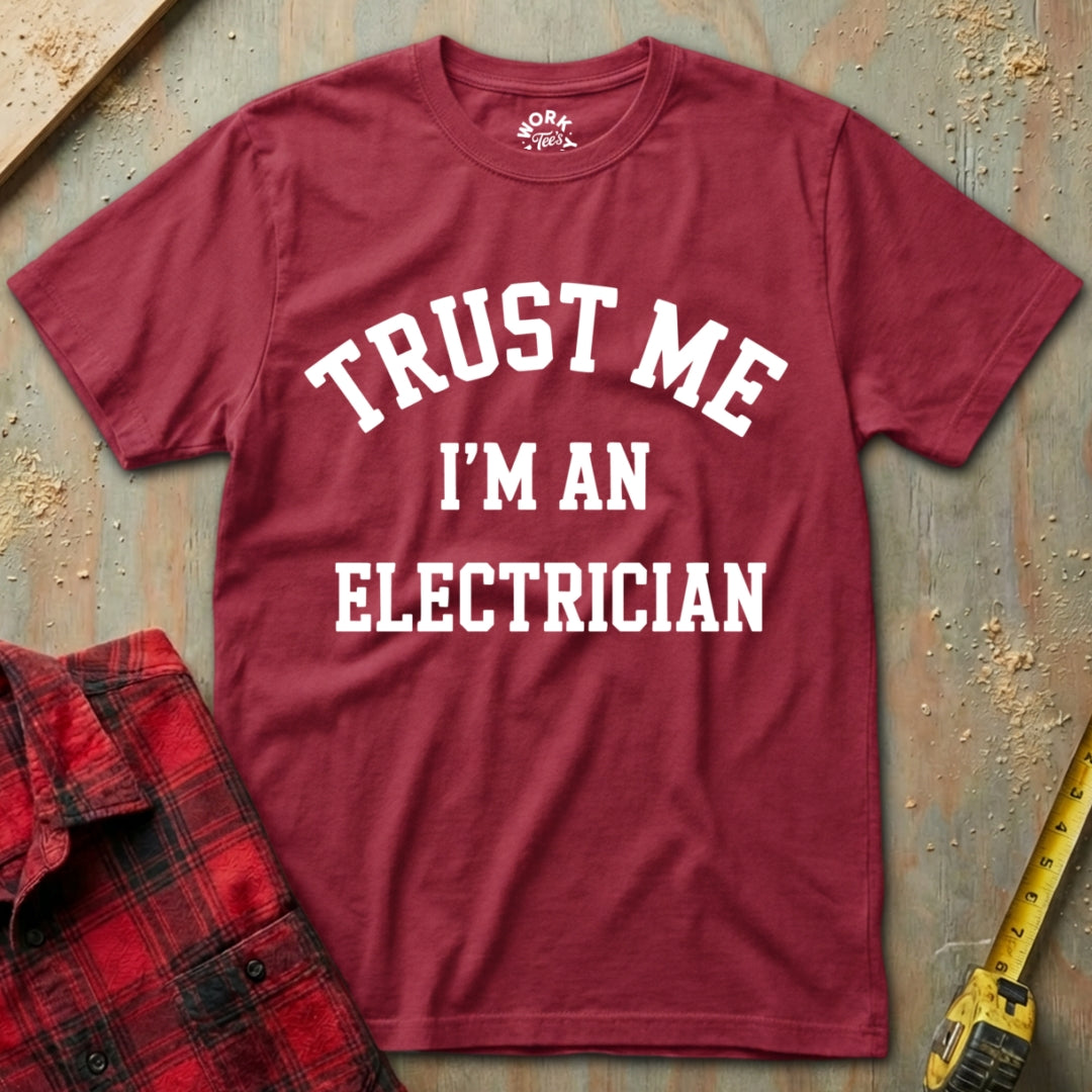 Red t-shirt with 'Trust Me I'm an Electrician' text on a textured surface with tools.