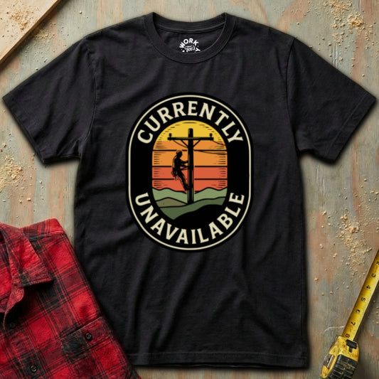 Black t-shirt with a graphic design of a person on a power line and 'Currently Unavailable' text, placed on a textured surface with a red shirt and yellow tool in the background.