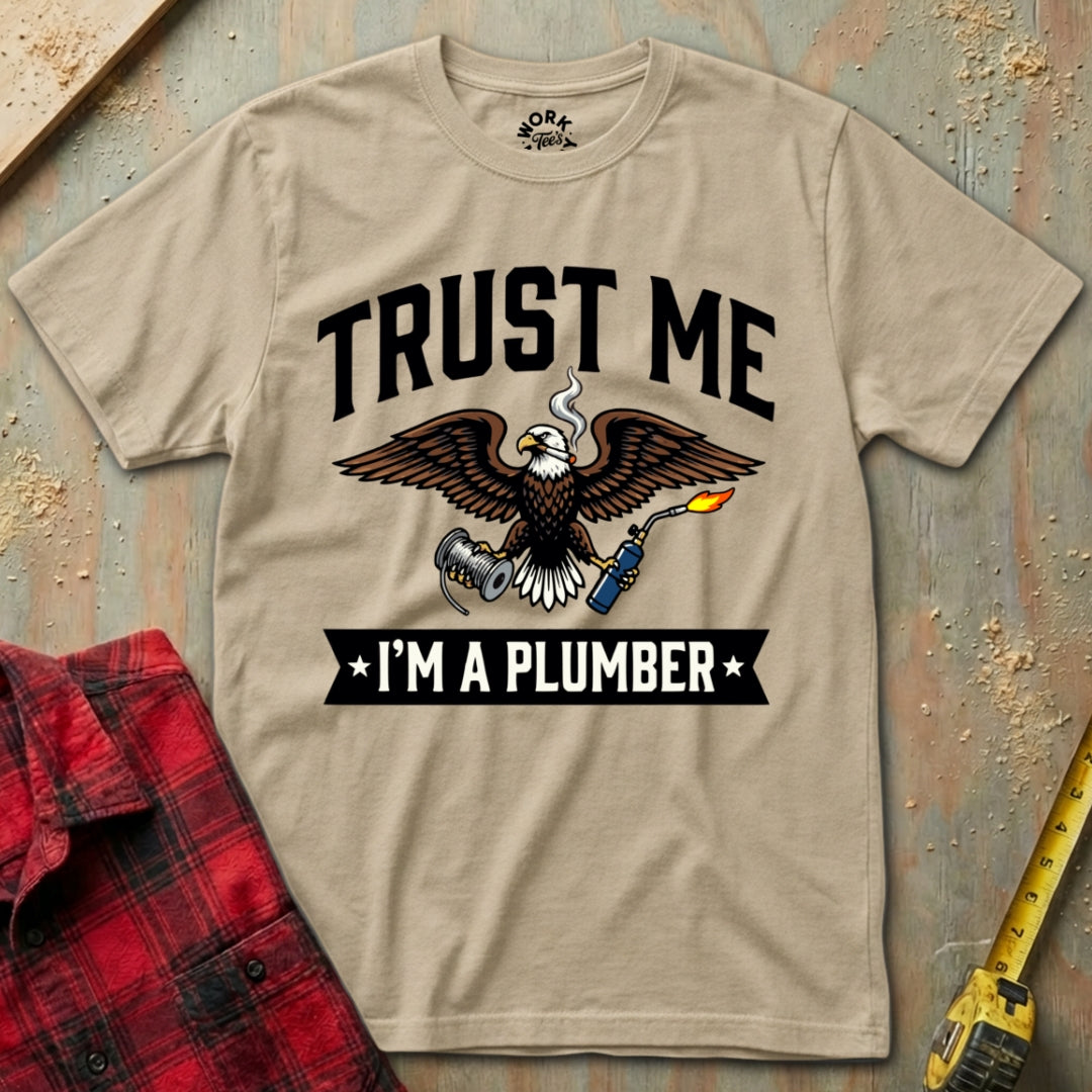 Beige t-shirt with eagle graphic and text 'Trust Me I'm a Plumber' on a textured surface with plaid shirt and tape measure.