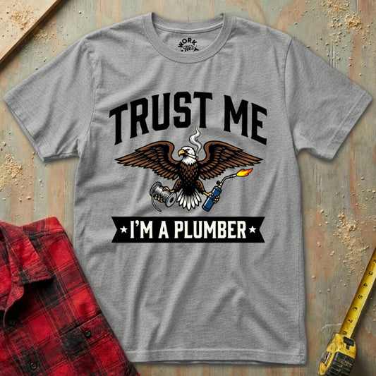 Gray t-shirt with an eagle graphic and text 'Trust Me I'm a Plumber' on a textured surface.