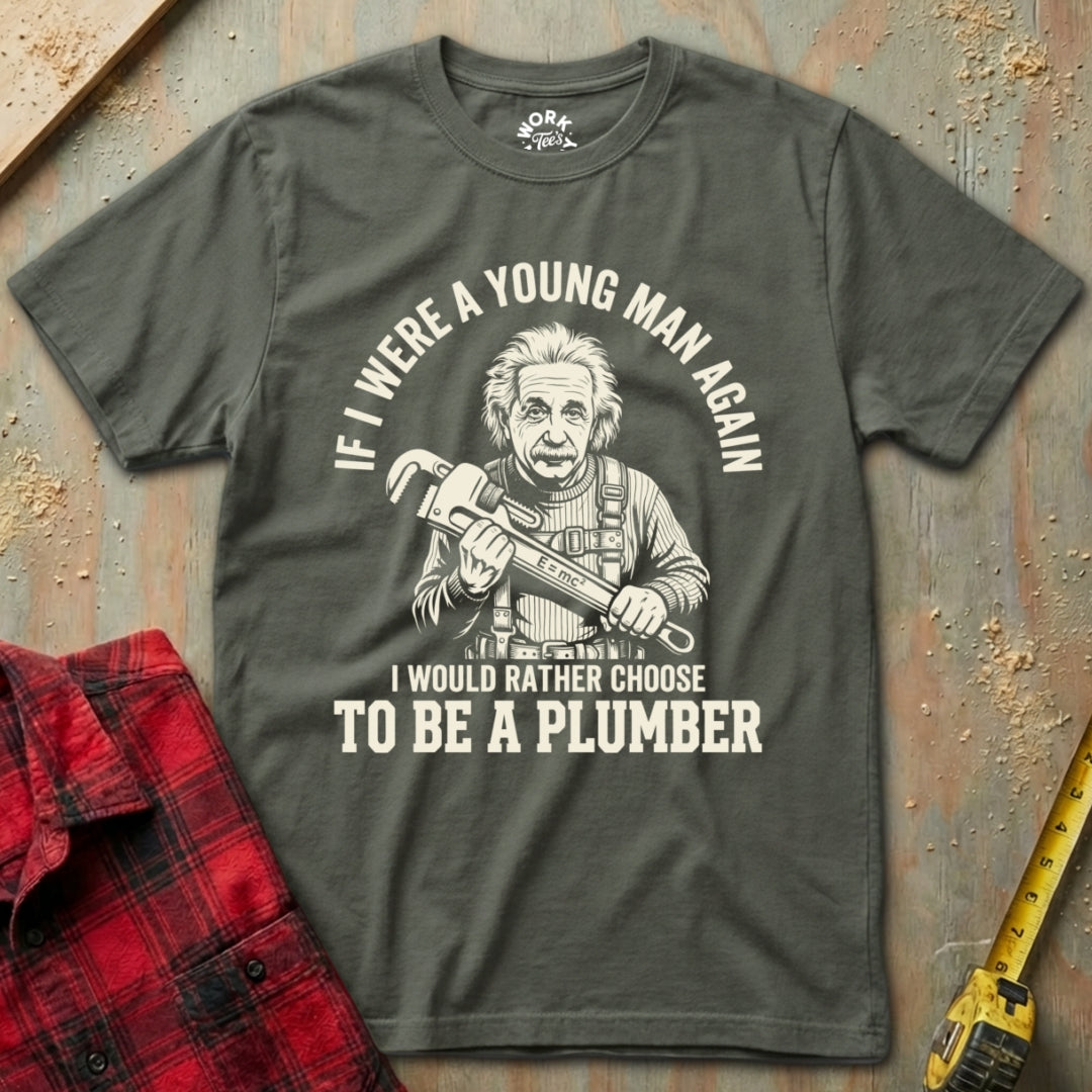 Gray t-shirt with a graphic and text about choosing to be a plumber, placed on a textured surface with a red flannel shirt and yellow tool in the background.