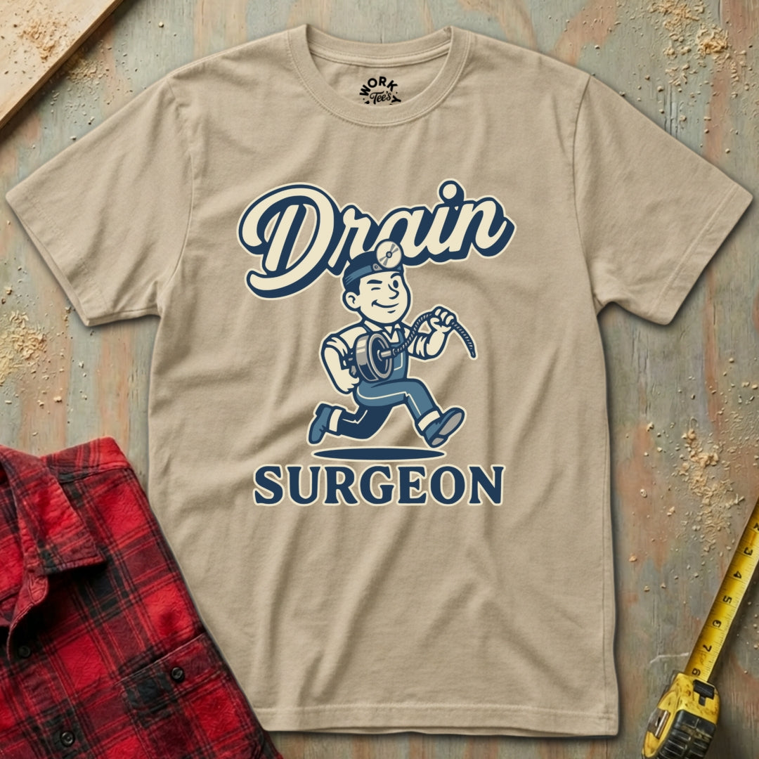 Beige t-shirt with 'Drain Surgeon' graphic design on a wooden surface with tools.