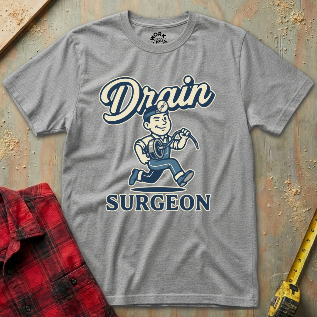 Gray t-shirt with 'Drain Surgeon' graphic design on a textured surface with plaid shirt and tape measure.