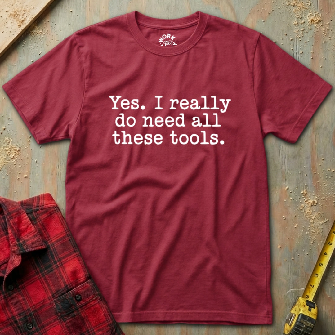 Red t-shirt with text 'Yes. I really do need all these tools.' on a textured surface with a plaid shirt and tape measure.