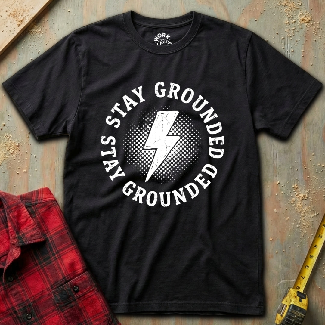 Black t-shirt with 'Stay Grounded' graphic on a textured surface with a red plaid shirt and yellow tool in the background.