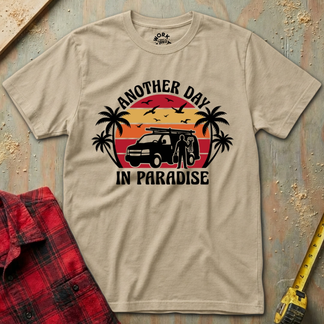 Beige t-shirt with 'Another Day in Paradise' graphic design on a wooden surface with a red plaid shirt and yellow tool in the background.