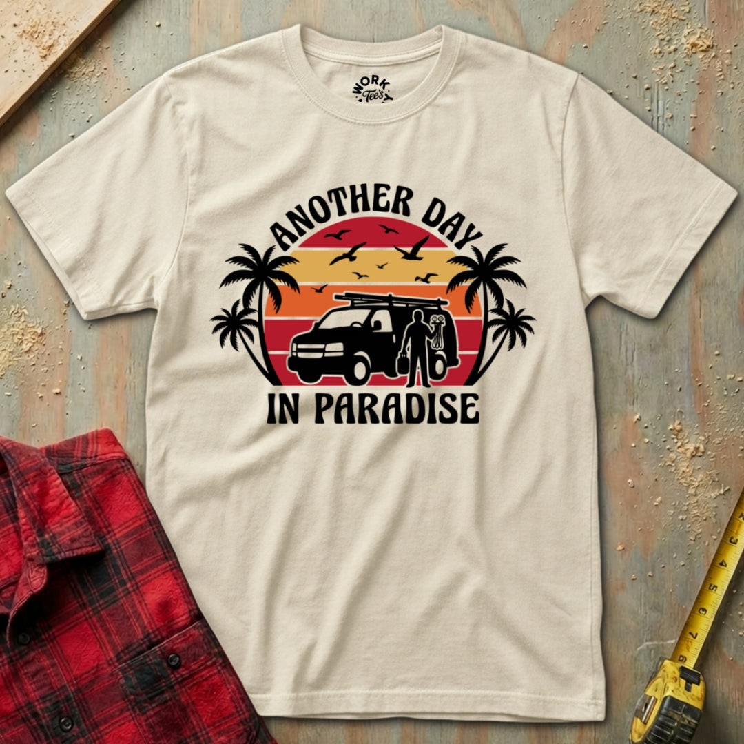 Beige t-shirt with 'Another Day in Paradise' graphic design on a textured surface with a red plaid shirt and yellow tool in the foreground.