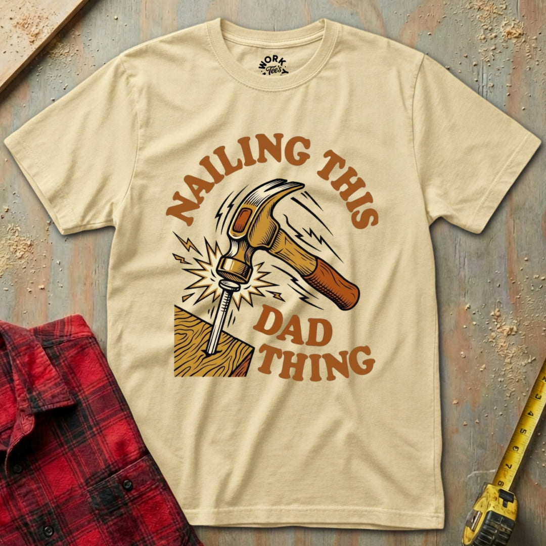 Beige t-shirt with a graphic design of a hammer and text 'Nailing this dad thing' on a rustic background.
