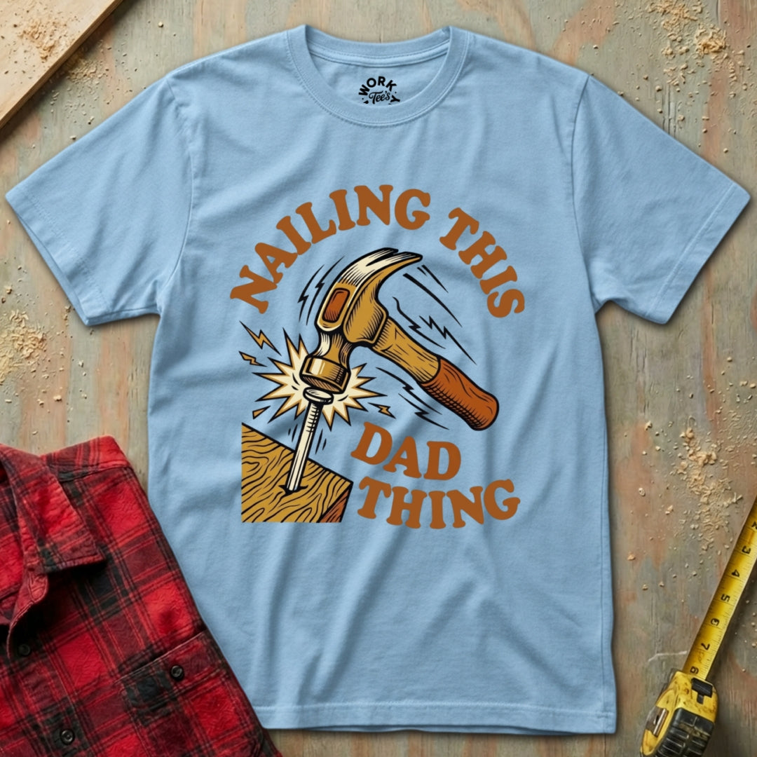 Light blue t-shirt with a graphic of a hammer and text 'Nailing this dad thing' on a wooden background.