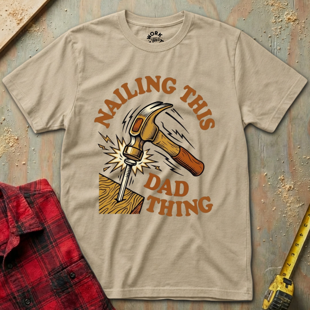 Beige t-shirt with a graphic design of a hammer and text 'Nailing this Dad Thing' on a wooden background.