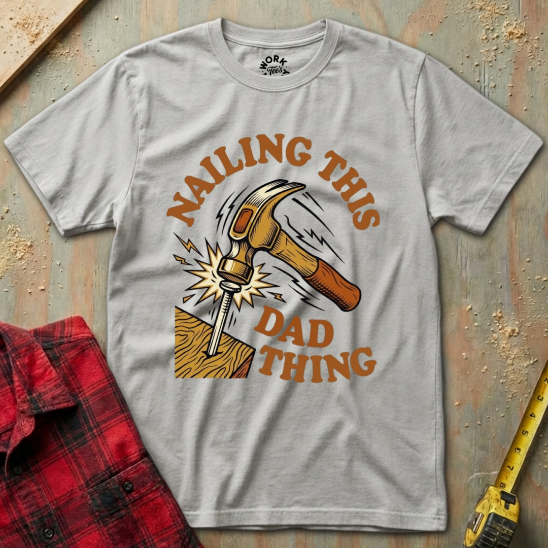 Gray t-shirt with a graphic design of a hammer and text 'Nailing this dad thing' on a wooden background.