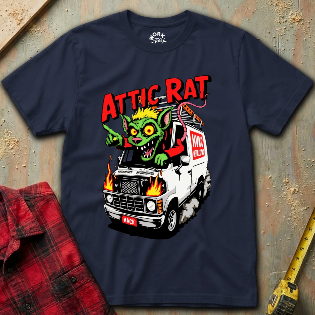 Navy blue t-shirt with a graphic of a rat on a truck and 'ATTIC RAT' text, placed on a textured surface with a red plaid shirt and yellow tool in the background.