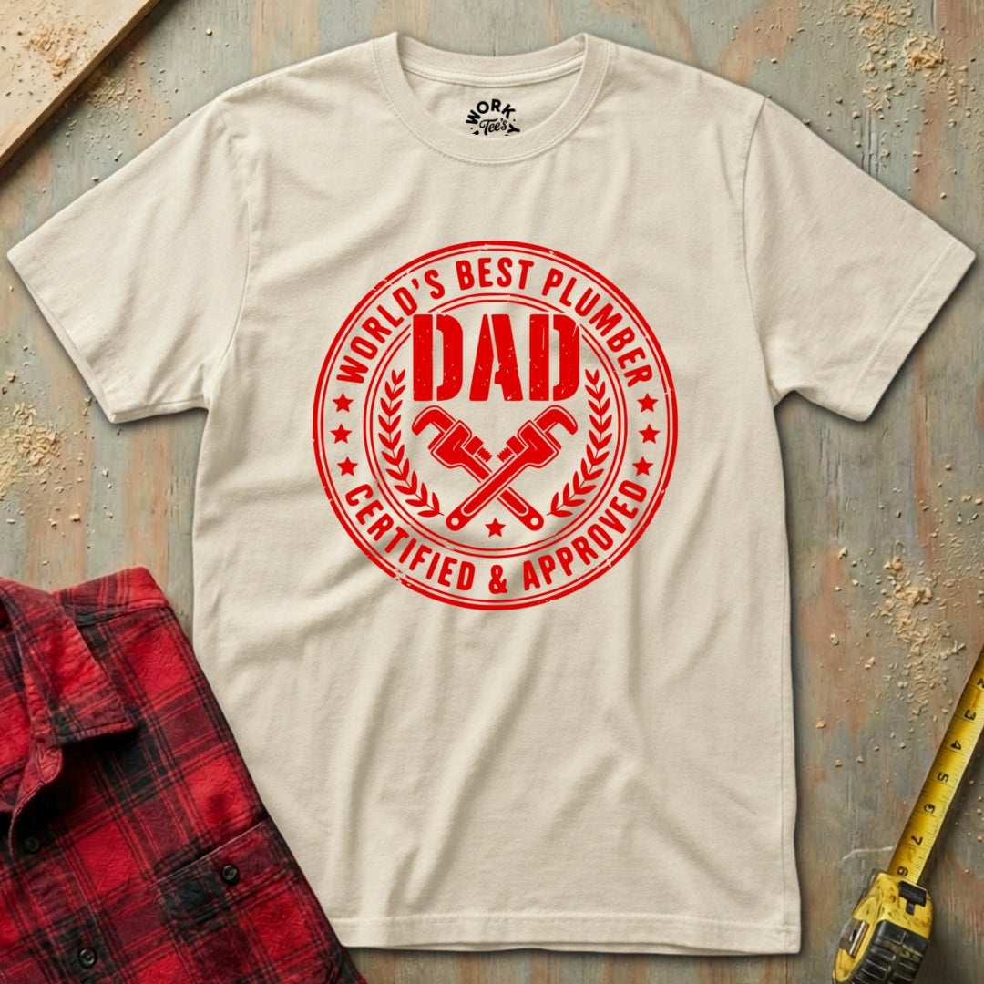 Beige t-shirt with red 'World's Best Plumber Dad' graphic on a textured surface with plaid shirt and tape measure.