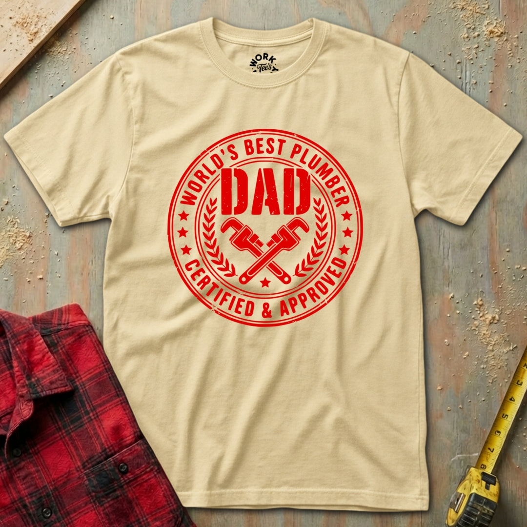 Beige t-shirt with red 'World's Best Plumber Dad' graphic on a textured surface with tools.
