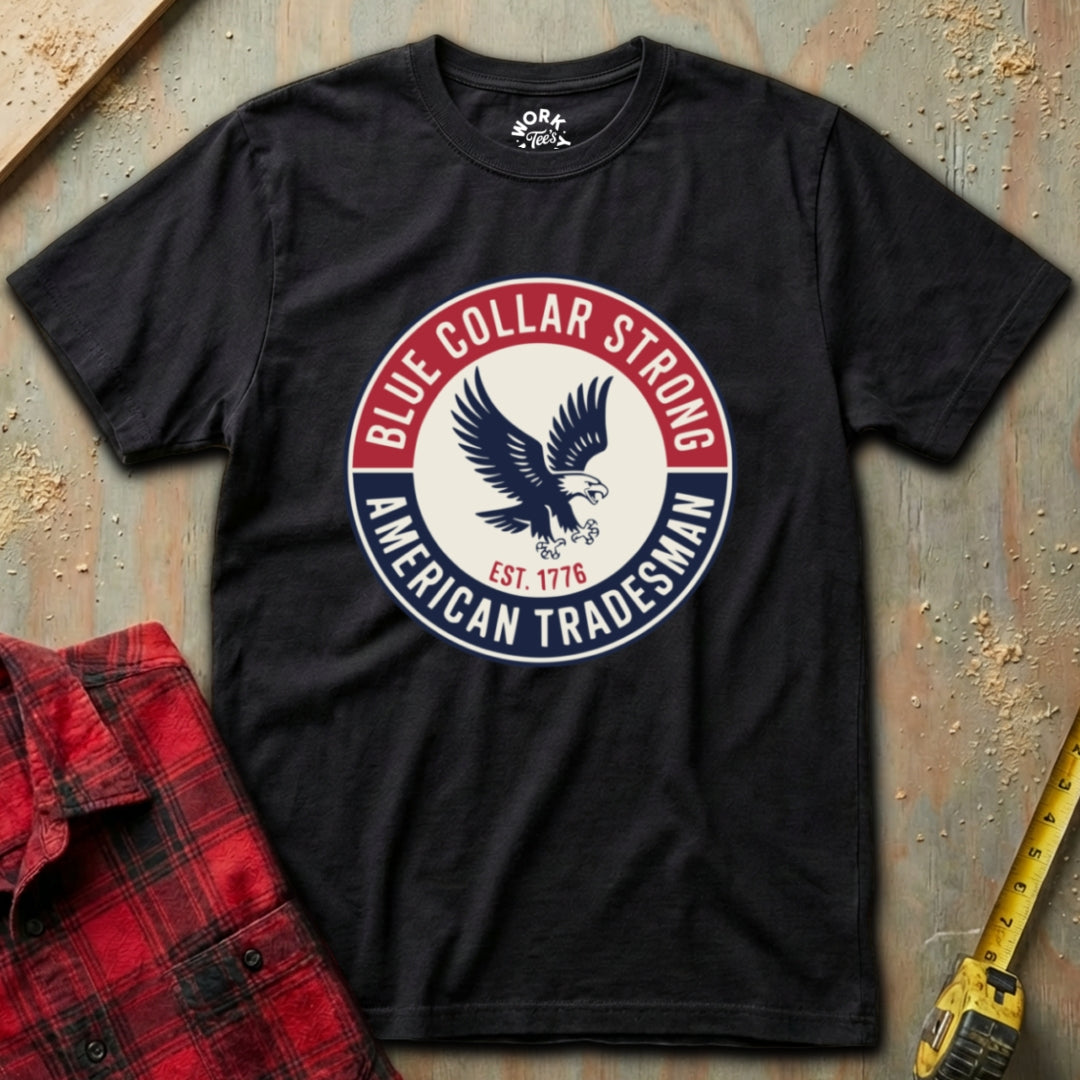 Black t-shirt with 'Blue Collar Strong American Tradesman' logo on a textured surface with a red plaid shirt and yellow tool in the background.