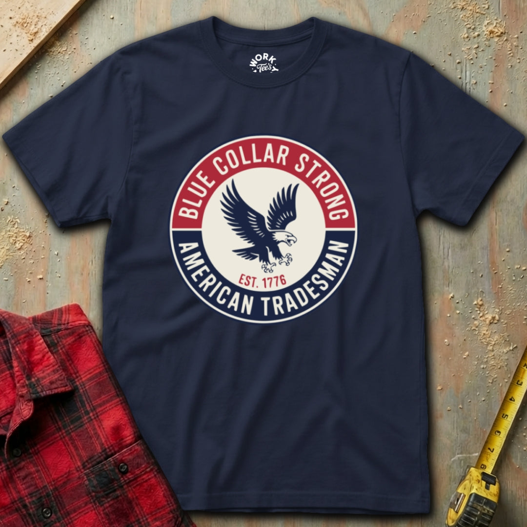 Navy blue t-shirt with 'Blue Collar Strong American Tradesman' graphic on a textured surface with plaid shirt and tape measure.