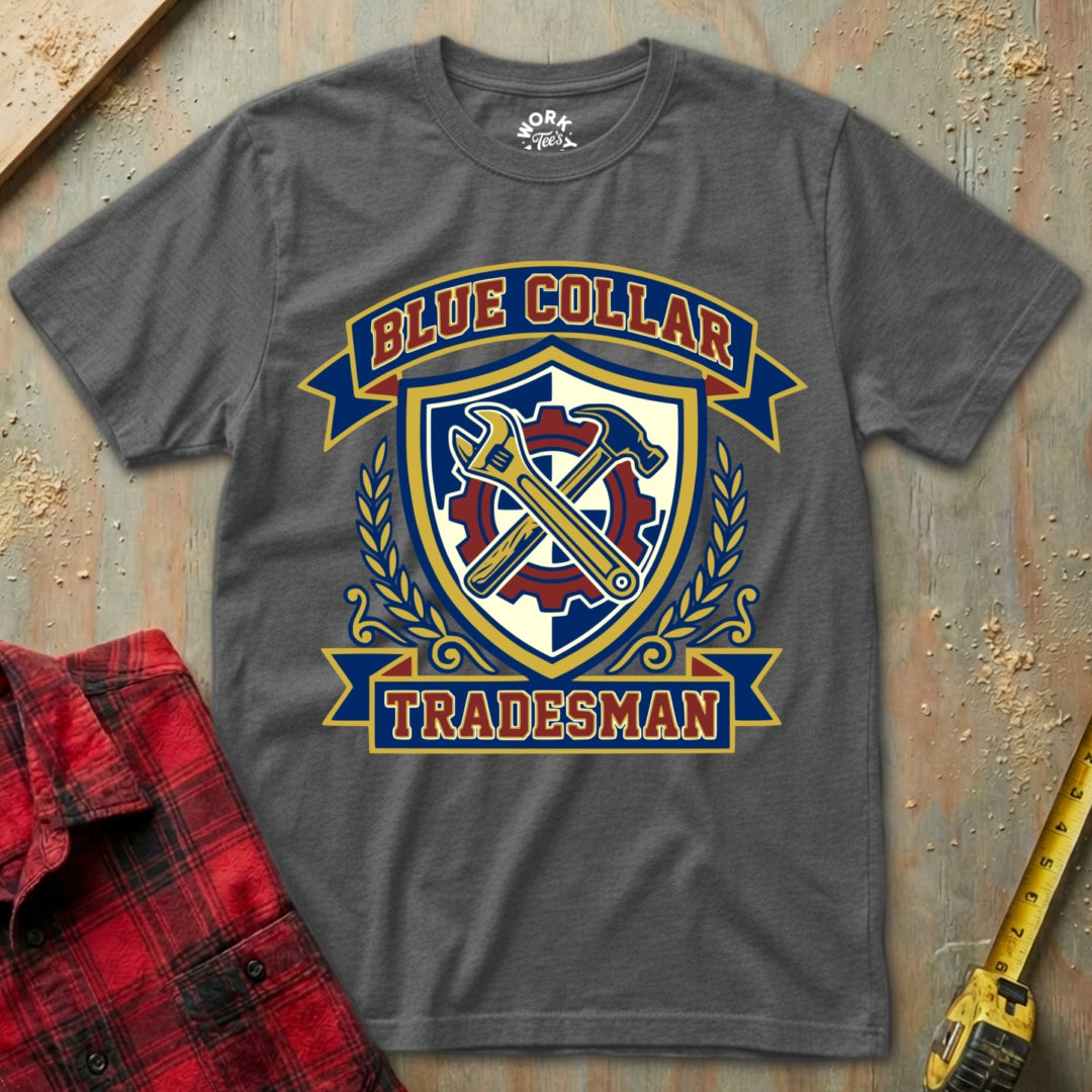 Gray t-shirt with 'Blue Collar Tradesman' graphic design on a textured surface with a red flannel shirt and yellow tool in the background.