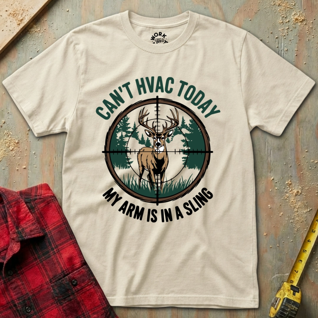 Beige t-shirt with a graphic of a deer and text 'Can't HVAC Today, My Arm is in a Sling' on a wooden surface with a red plaid shirt and yellow tool.