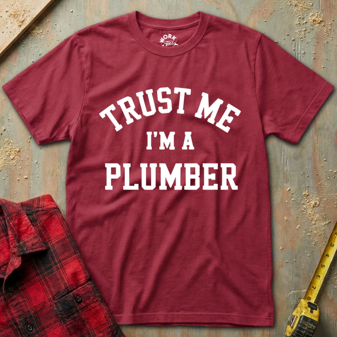 Red t-shirt with 'Trust Me I'm a Plumber' text on a textured surface with tools.
