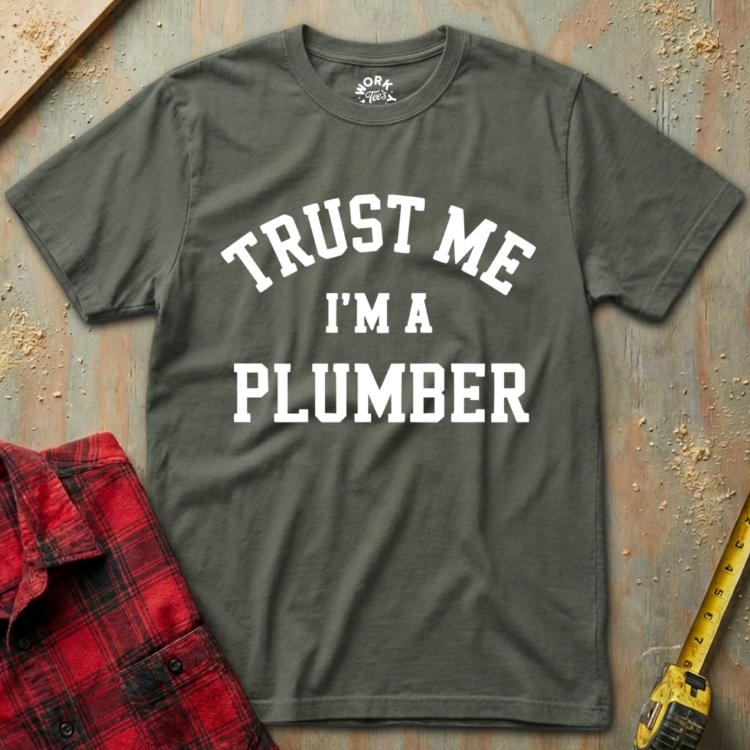 Gray t-shirt with 'Trust Me I'm a Plumber' text on a textured surface with a red plaid shirt and yellow tool in the background.