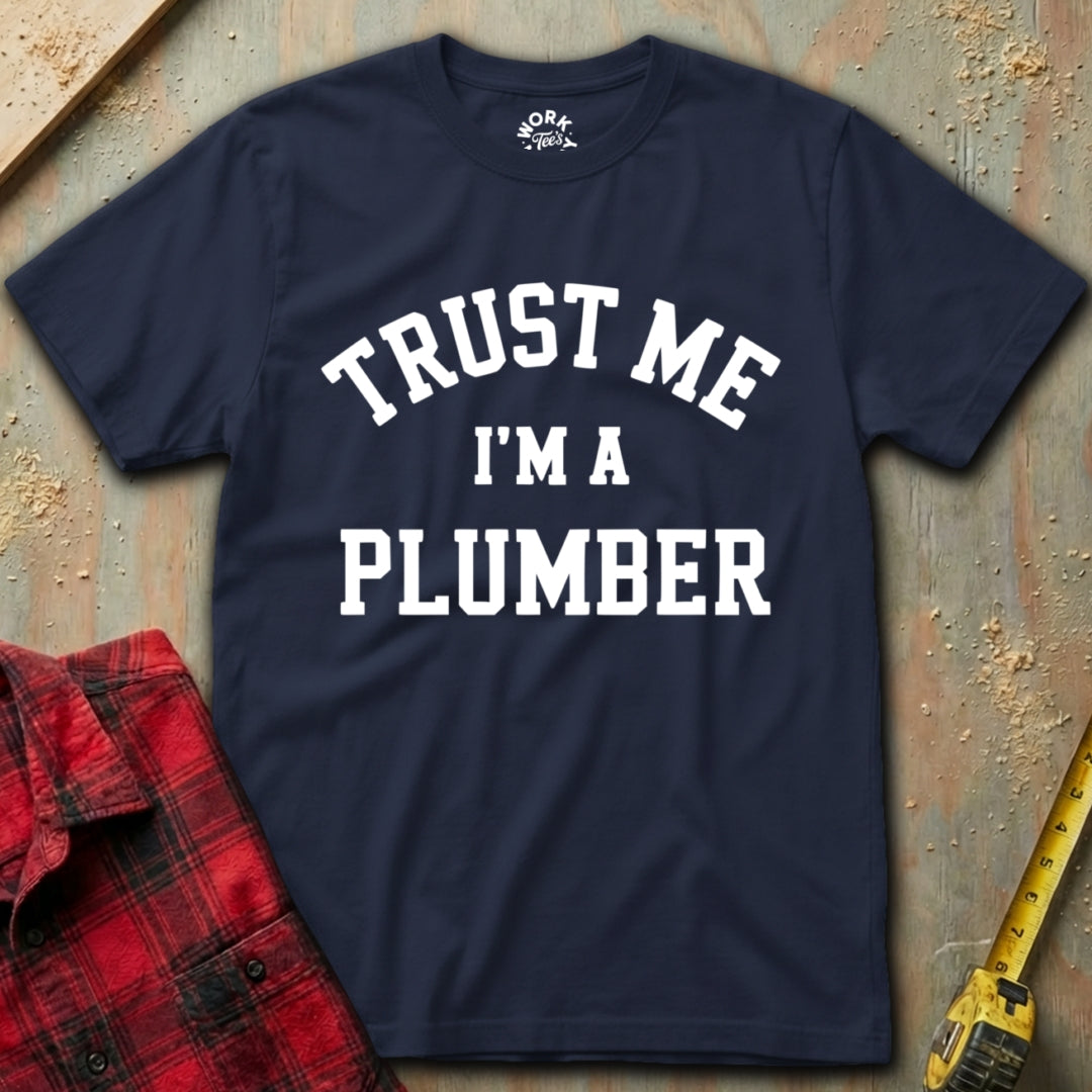 Navy blue t-shirt with 'Trust Me I'm a Plumber' text on a textured surface with tools.