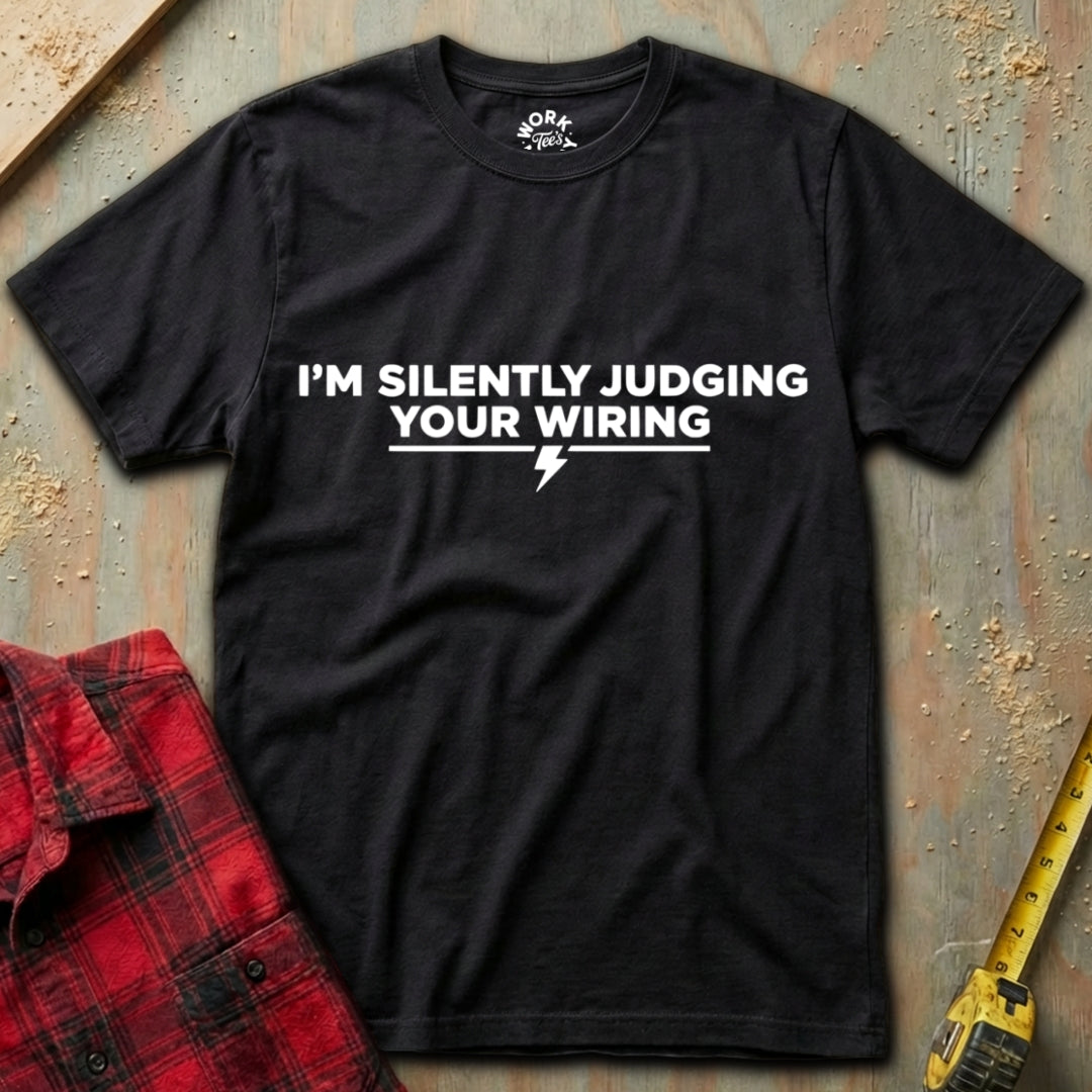 Black t-shirt with humorous text design on a textured surface with a red plaid shirt and yellow tool in the background.