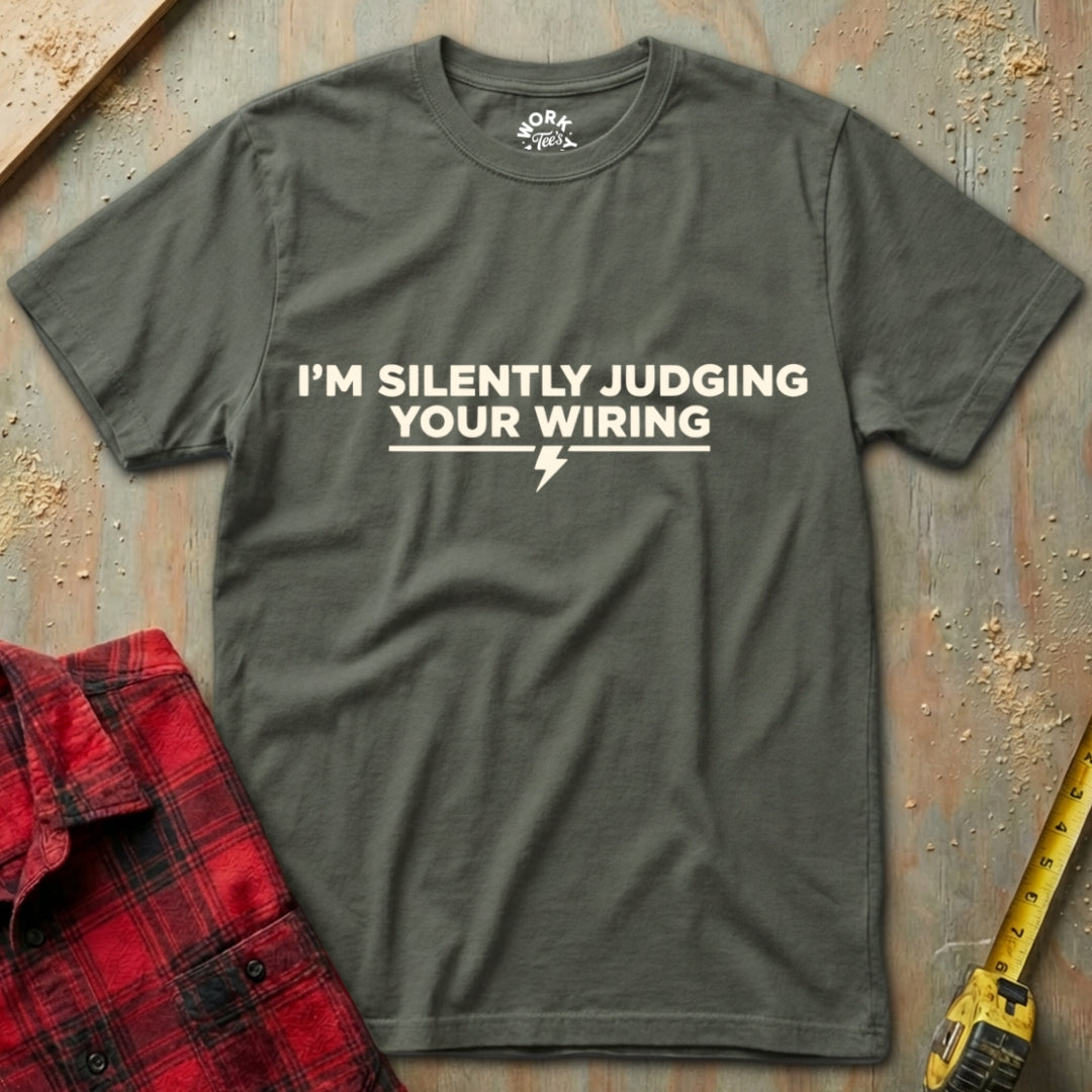 Gray t-shirt with humorous text design on a textured surface with a red plaid shirt and yellow tool in the background.