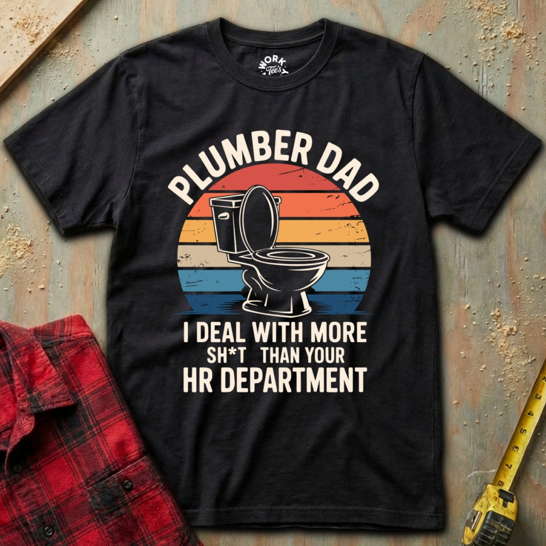 Black t-shirt with 'Plumber Dad' graphic and text on a textured surface with plaid shirt and tape measure.