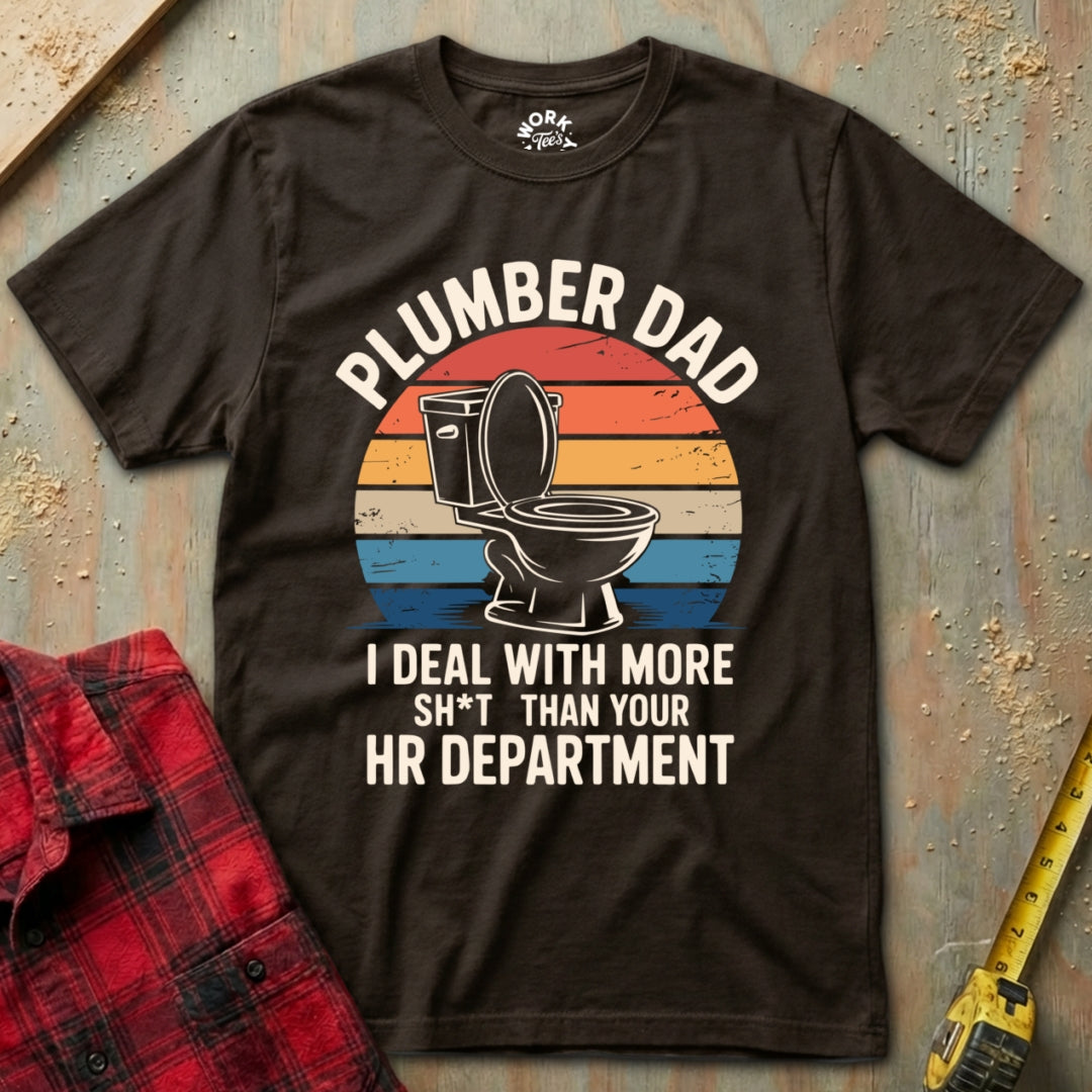 Brown t-shirt with 'Plumber Dad' graphic and text on a textured surface with plaid shirt and tape measure.