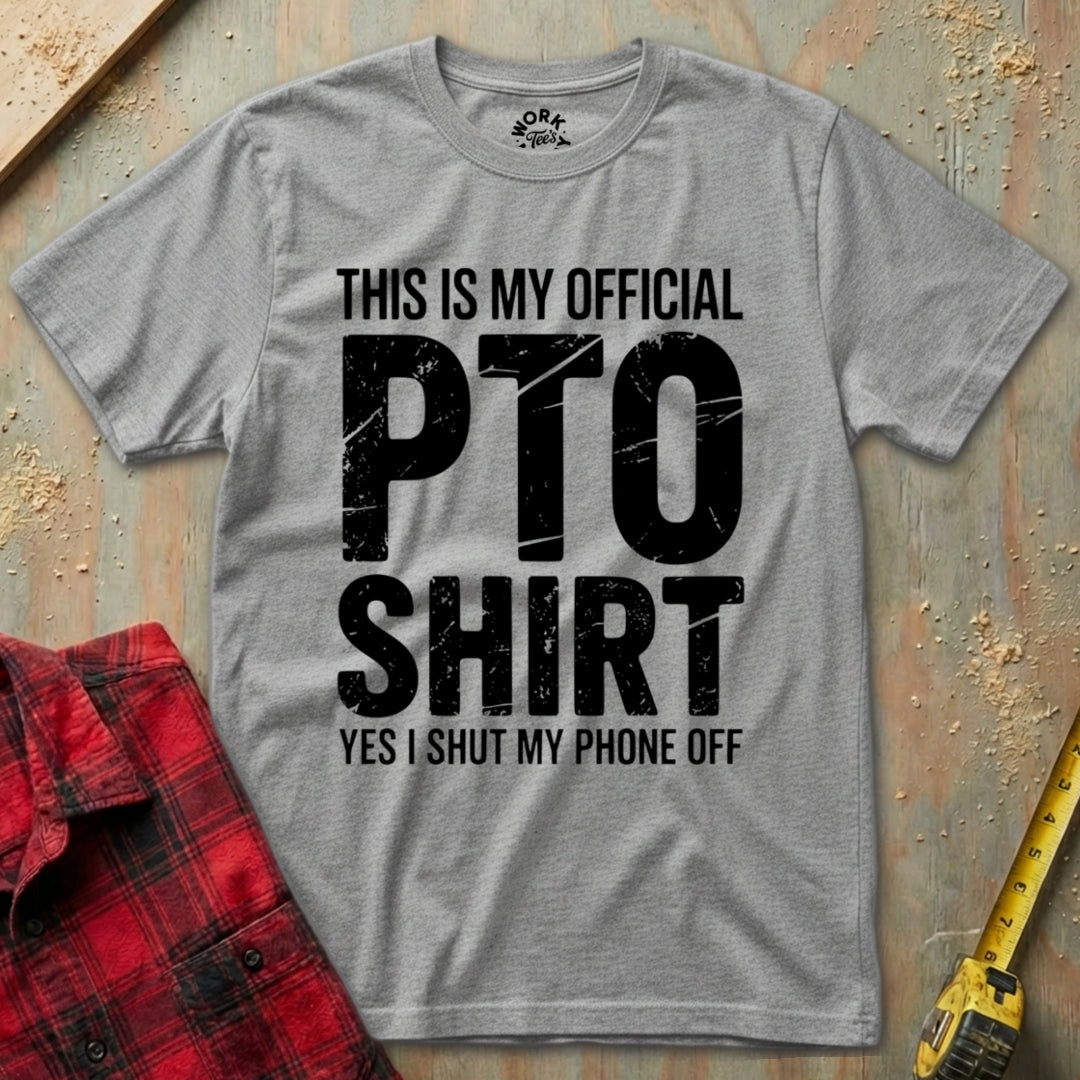Gray t-shirt with 'This is my official PTO shirt' text on a textured surface with a red plaid shirt and yellow tool in the background.