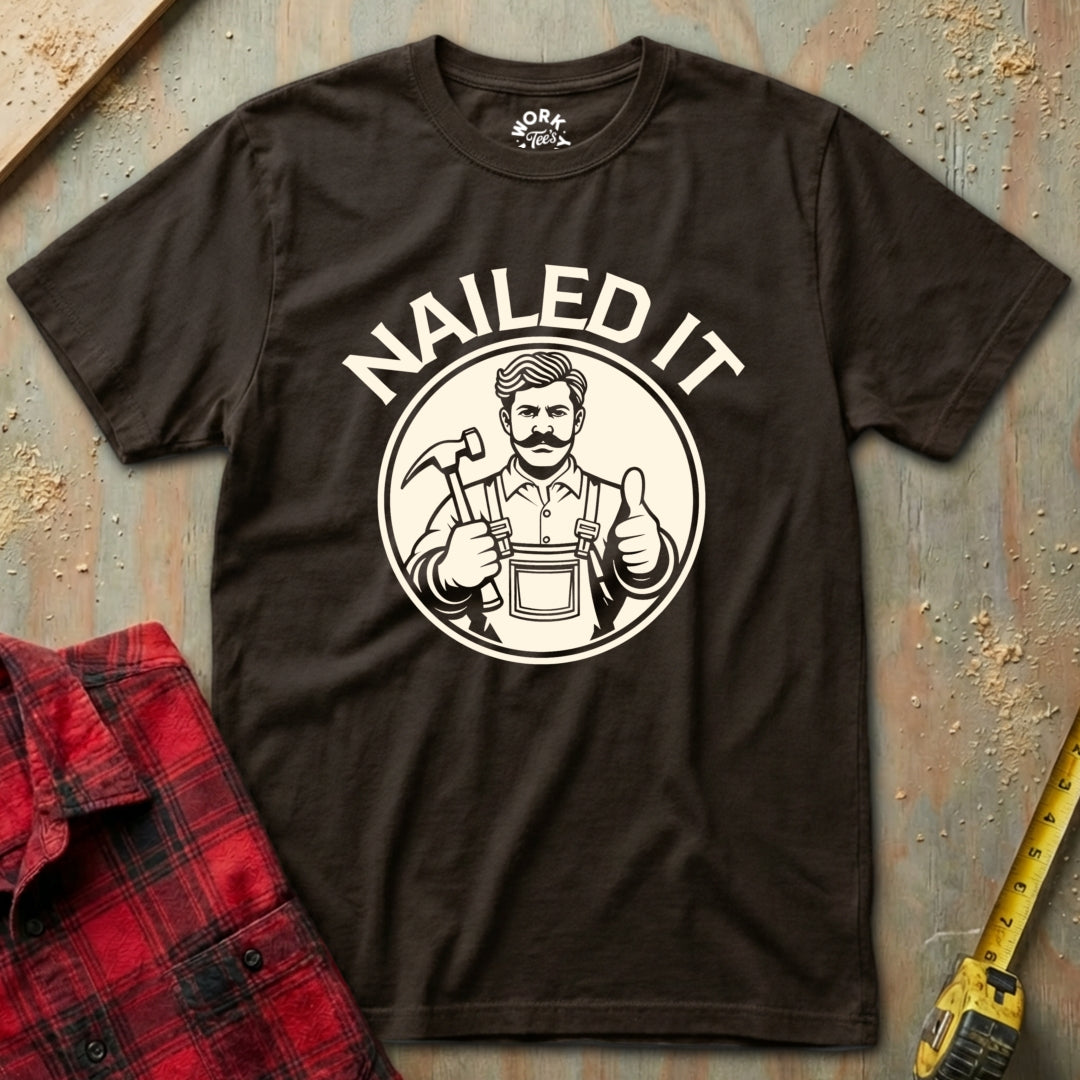 Brown t-shirt with 'NAILED IT' graphic on a textured surface with a red plaid shirt and yellow tape measure.