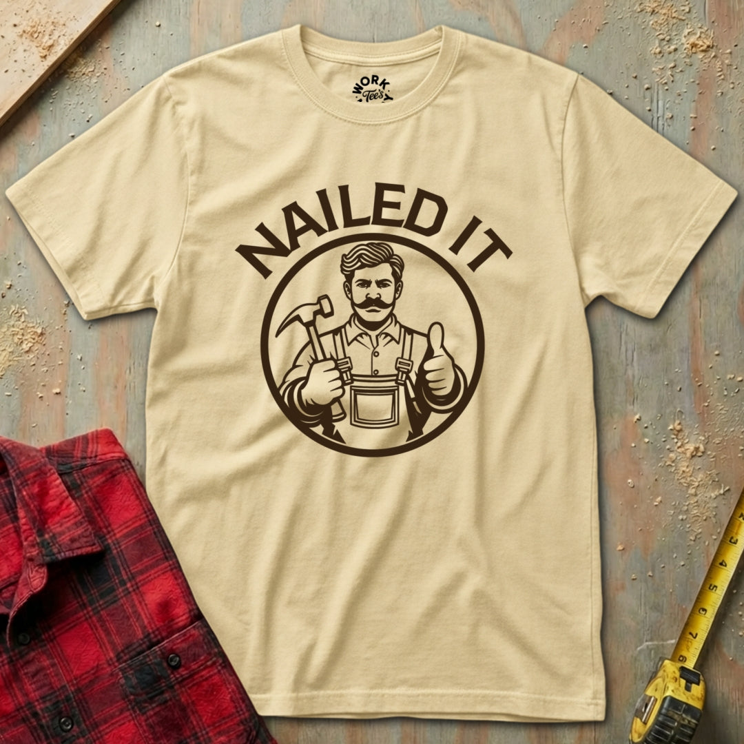Beige t-shirt with 'NAILED IT' graphic on a textured surface with plaid shirt and tape measure.