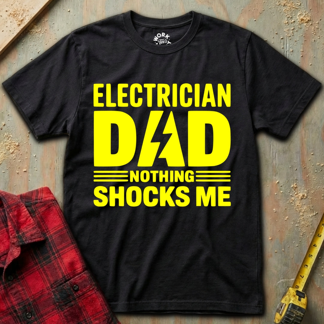 Black t-shirt with yellow text 'Electrician Dad Nothing Shocks Me' on a textured surface with plaid shirt and drill.