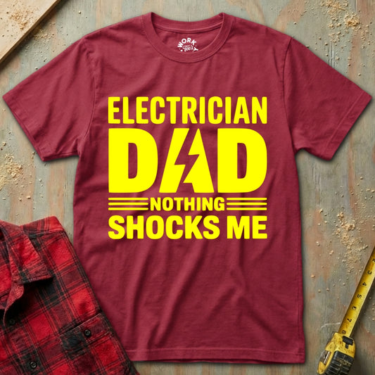 Maroon t-shirt with yellow text 'Electrician Dad Nothing Shocks Me' on a textured surface.