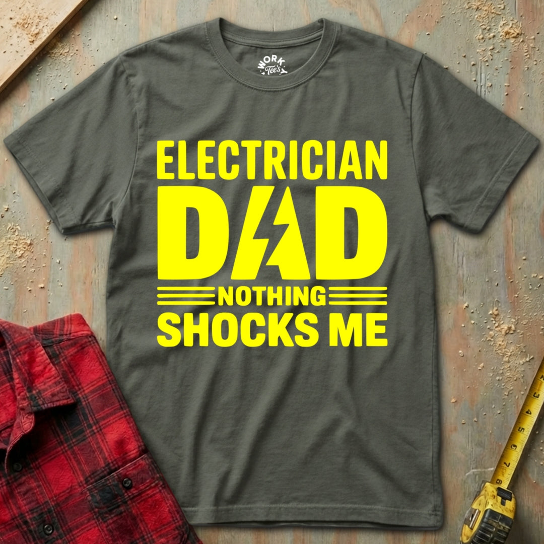 Gray t-shirt with yellow text 'Electrician Dad Nothing Shocks Me' on a textured surface with a red plaid shirt and yellow tool.