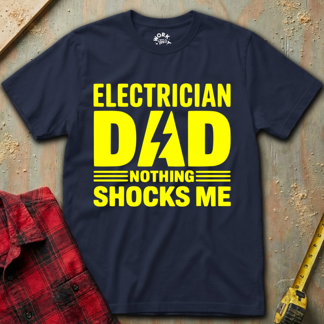 Navy blue t-shirt with yellow text 'Electrician Dad Nothing Shocks Me' on a textured surface with a red plaid shirt and yellow tool.