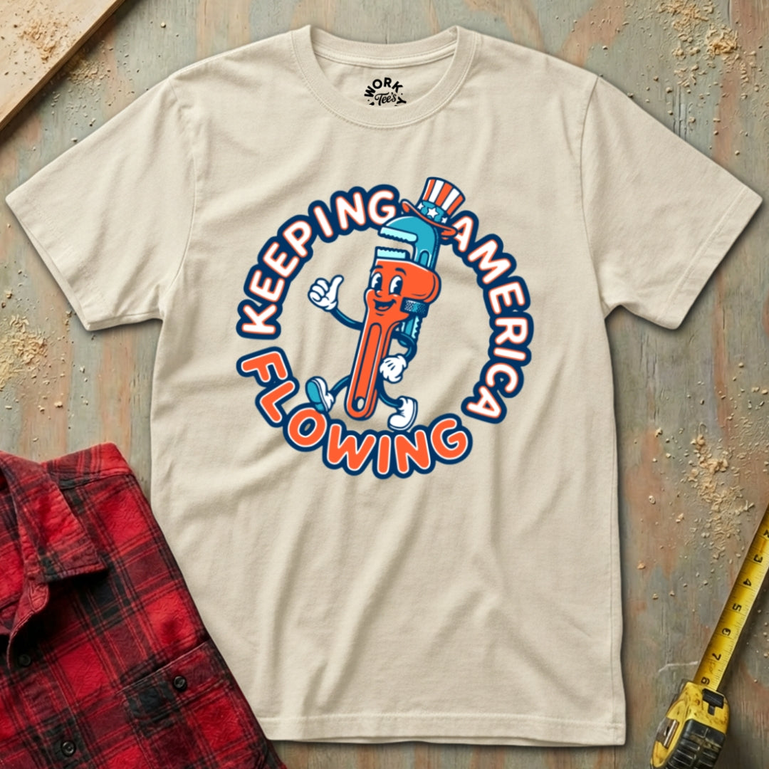 Beige t-shirt with a cartoon faucet graphic and text 'Keeping America Flowing' on a textured surface.
