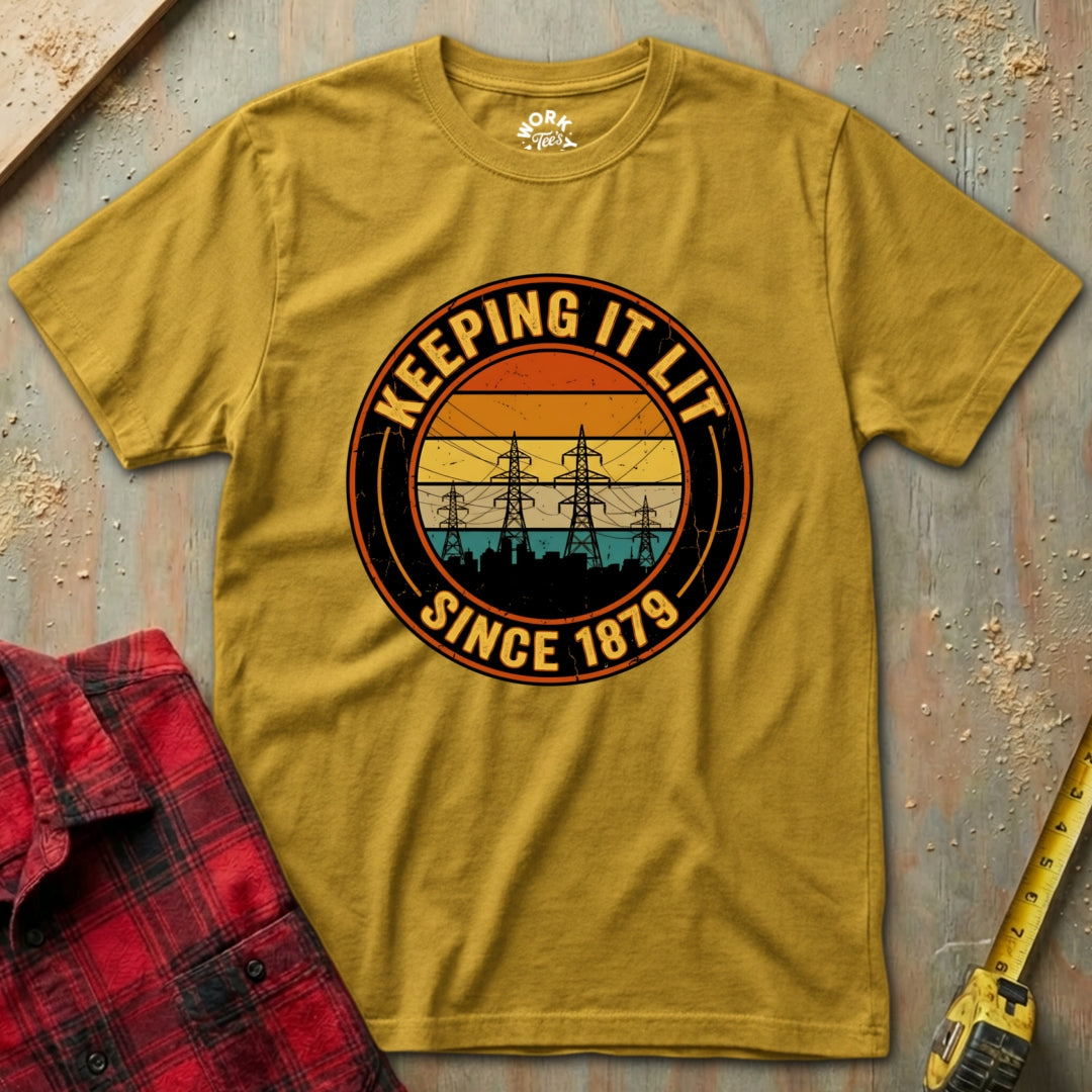 Yellow t-shirt with 'Keeping It Lit Since 1879' design on a textured surface with a red plaid shirt and yellow tool.