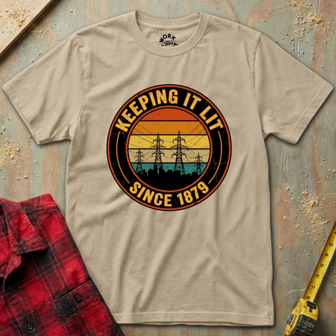 Beige t-shirt with 'Keeping It Lit Since 1879' graphic design on a textured surface with plaid shirt and tape measure.